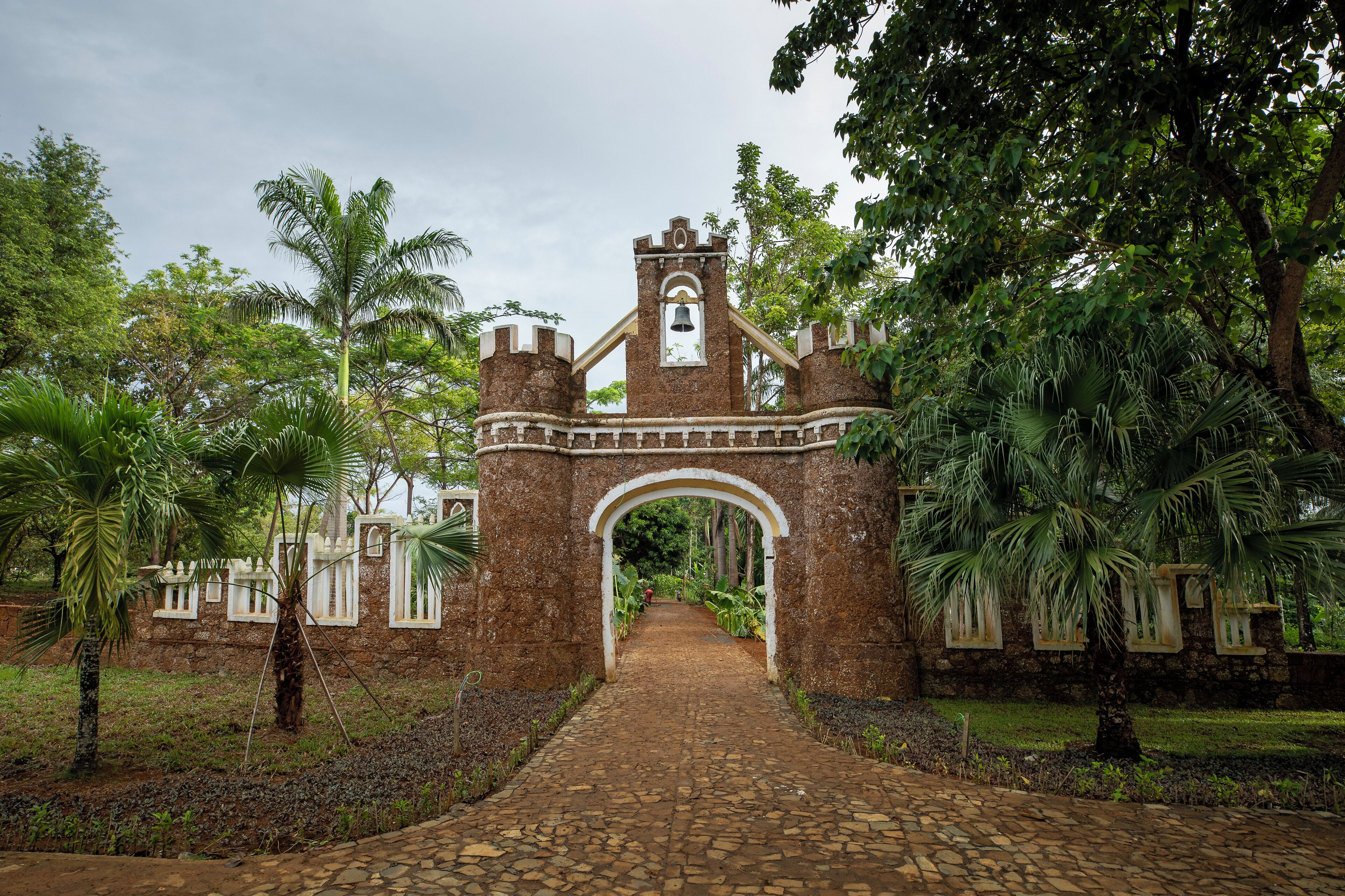 A stone archway leading to a building with a bell tower, surrounded by lush greenery and a cobblestone path.