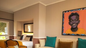 A living room with a sofa, armchair, and coffee table. There is a painting on the wall and a window with a view of trees.