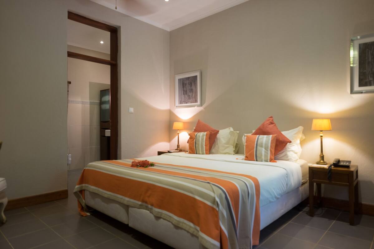A hotel room with a bed, two bedside lamps, a framed picture on the wall, and a door leading to another room.