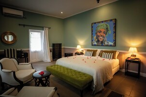 Habitación de hotel con cama, un reposapiés verde, una silla, un escritorio, una lámpara y un cuadro en la pared.