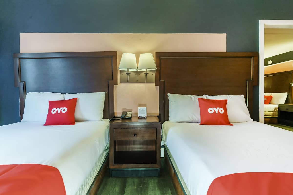Habitación doble con dos camas, cada una con cabecero de madera, ropa de cama blanca y almohadas rojas con la palabra 'OYO'. Hay mesitas de noche con lámparas y un cajón pequeño.