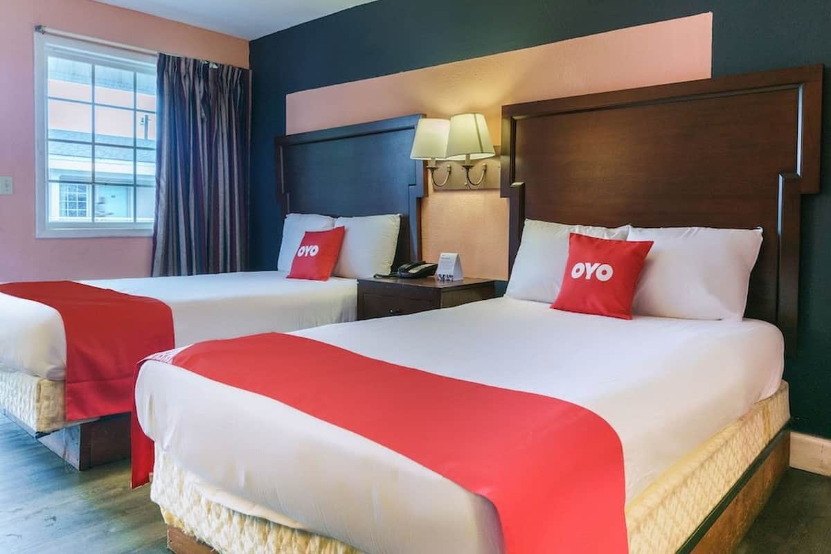 Habitación de hotel con dos camas, cada una con almohada roja con la marca OYO, una mesita de noche con lámpara y una ventana con cortinas.