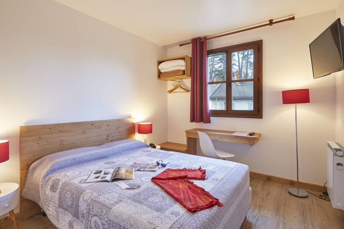 Un dormitorio con cabecera de madera, cama con edredón estampado, escritorio con silla, televisor montado en la pared y ventana con cortinas.