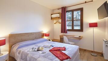 Une chambre à coucher avec une tête de lit en bois, un lit recouvert d’une couvre-lit à motifs, un bureau avec une chaise, un téléviseur fixé au mur et une fenêtre avec des rideaux.