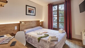 A hotel room with a wooden headboard, a bed with a patterned bedspread, a framed picture on the wall, a window with red curtains, and a television on the wall. - Cit’Hôtel Le National (Cluses)