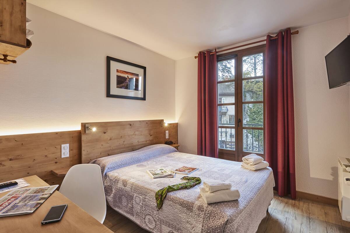 Una habitación de hotel con cabecera de madera, una cama con colcha estampada, un cuadro enmarcado en la pared, una ventana con cortinas rojas y un televisor en la pared.