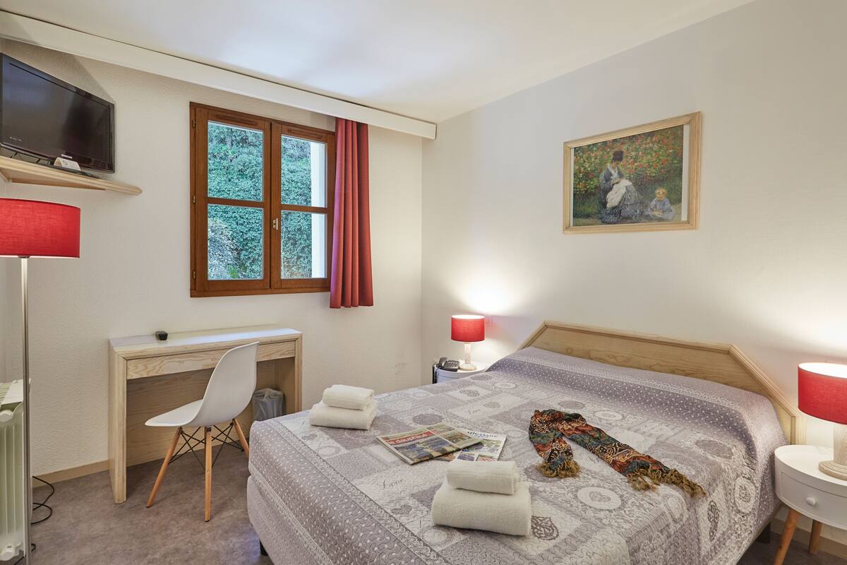 Un dormitorio con cama, escritorio, silla, televisor, ventana con cortinas, un cuadro y lámpara.