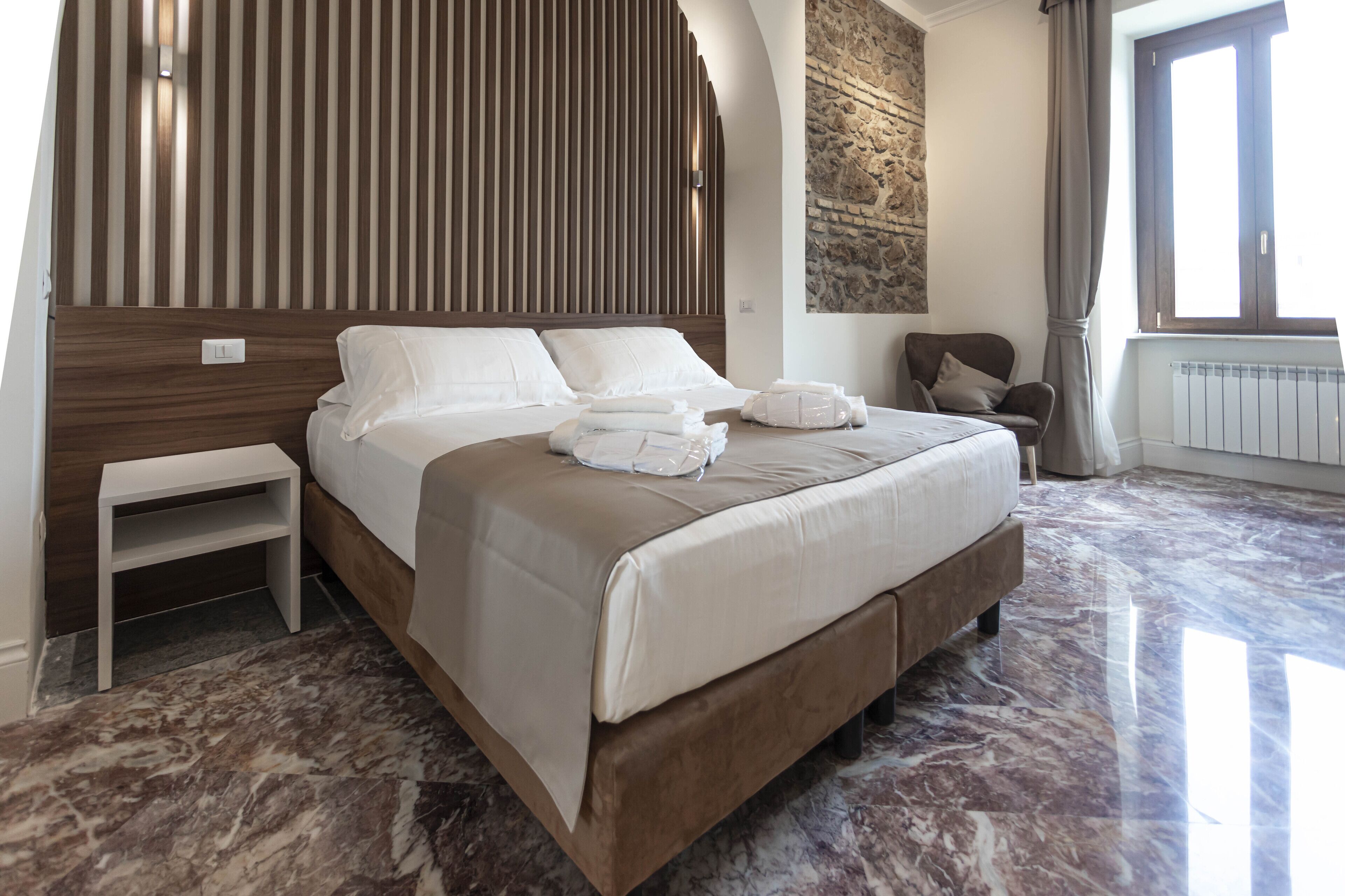 A modern hotel room with a large bed, a small bedside table, a stone accent wall, and a view of the outdoors.