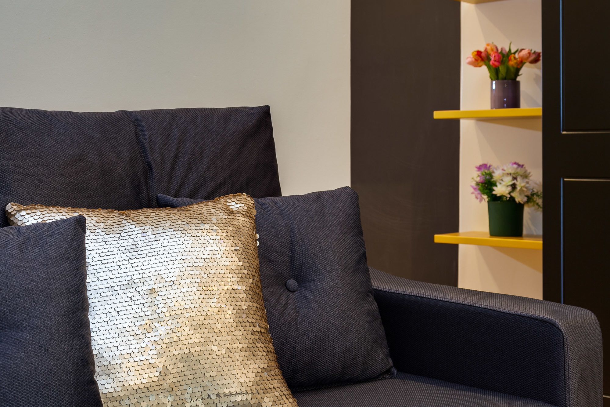 A dark-colored sofa with a sequined pillow and a cushion. There are two yellow shelves with flower arrangements.