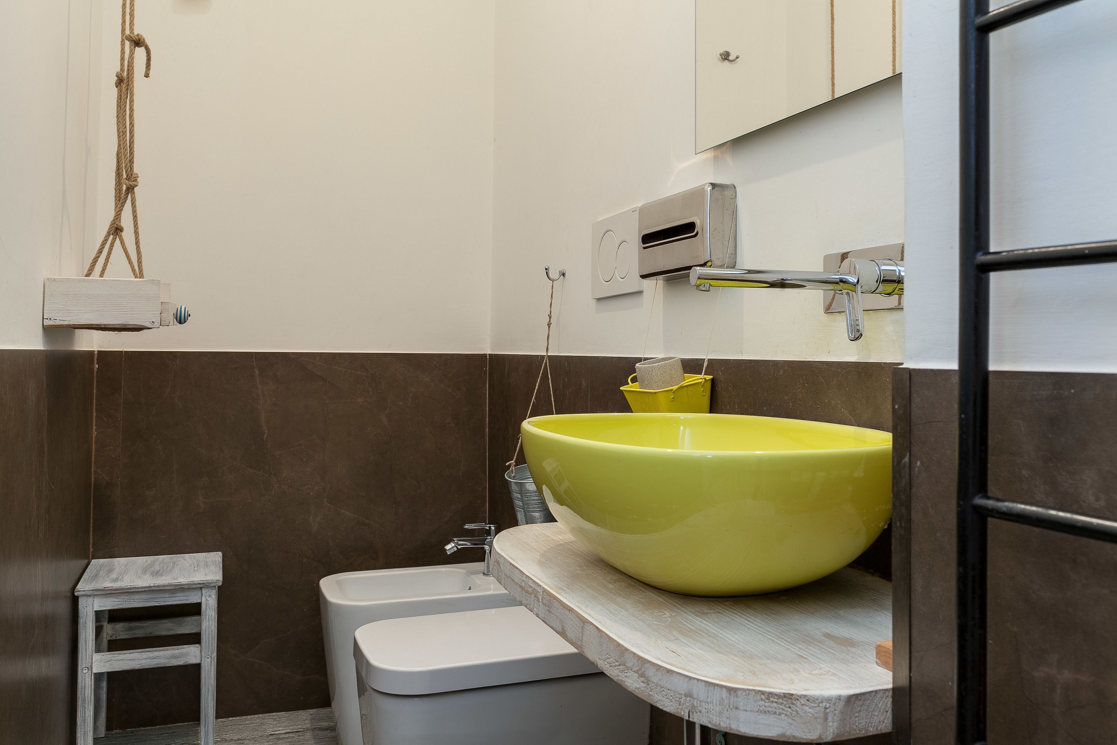 Une salle de bain avec un lavabo jaune, un petit tabouret blanc, des toilettes blanches et un espace douche.