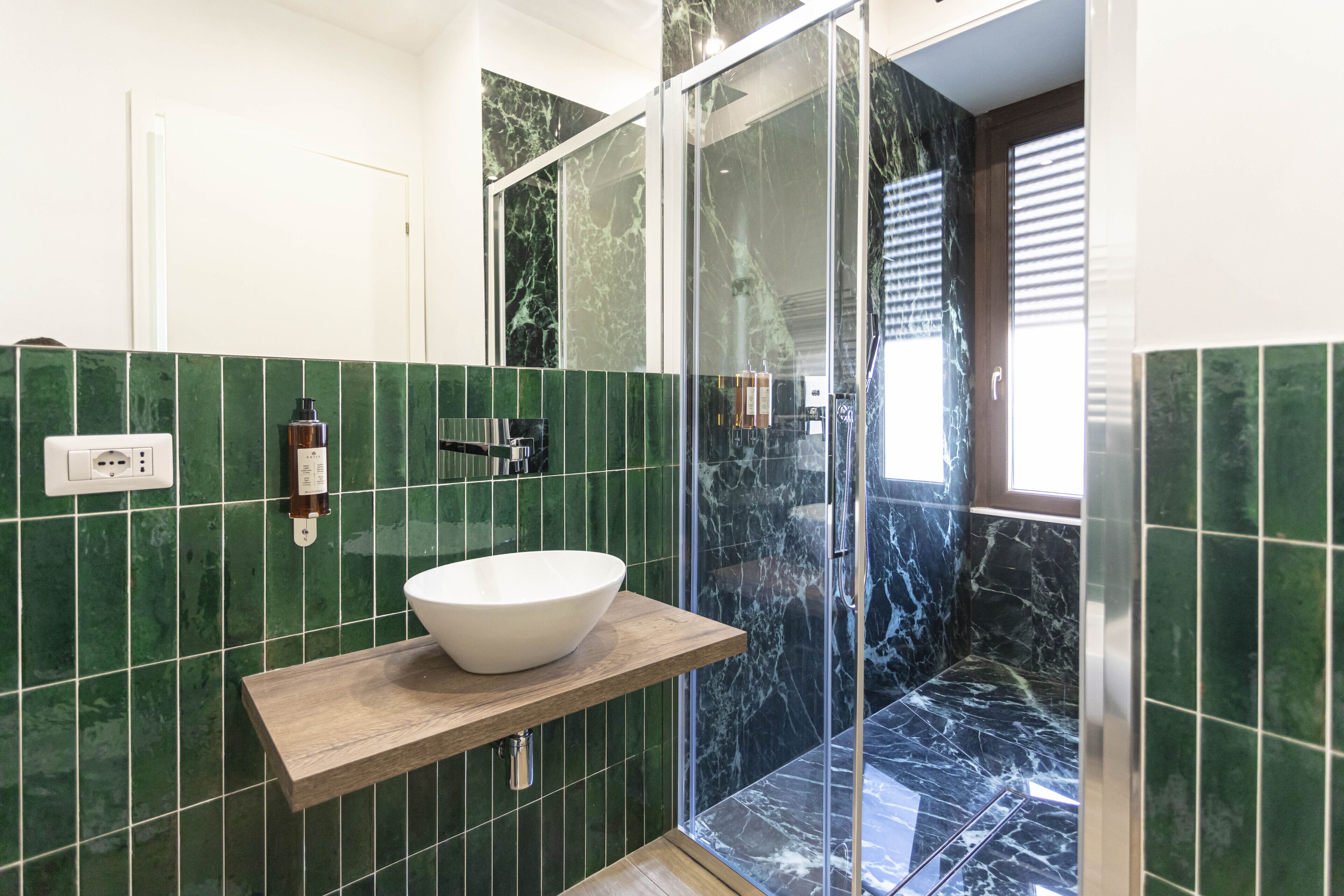A bathroom with a white basin on a wooden shelf, green marble tiles, a glass shower enclosure, and a door leading to another room.