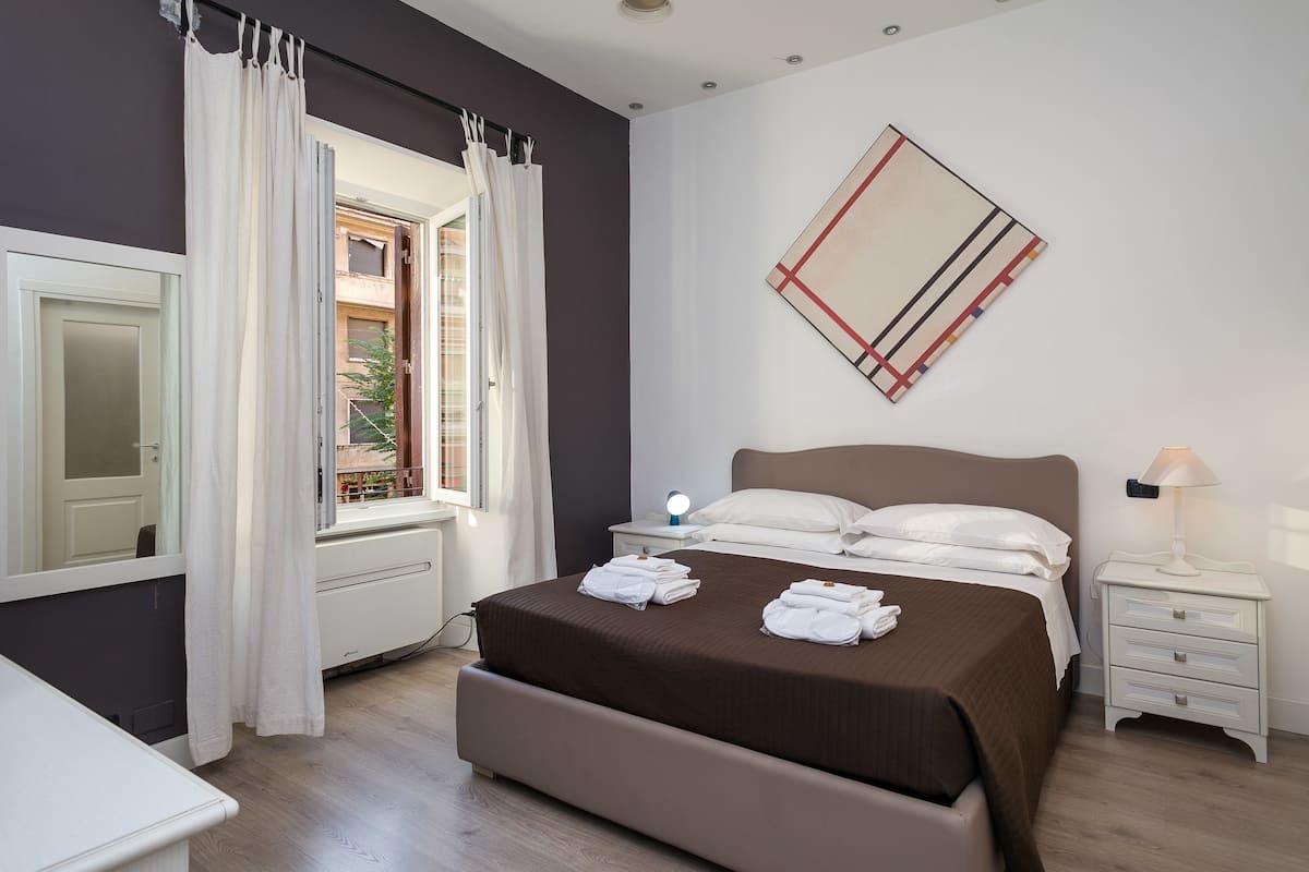 A bedroom with a large bed, a bedside table with a lamp, a mirror, and a window with curtains.