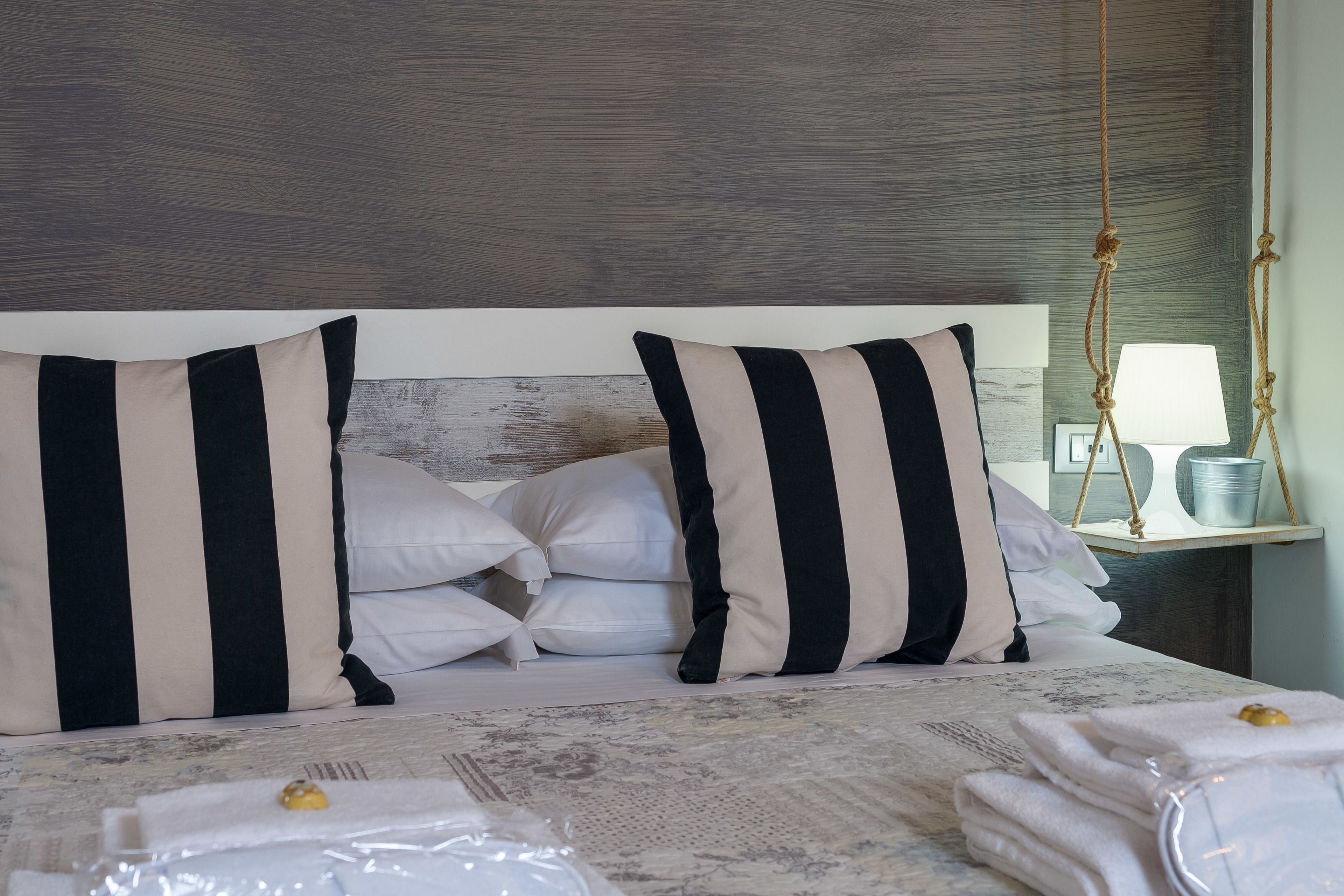 Una cama bien hecha con almohadas a rayas, una lámpara de noche y toallas dobladas ordenadamente sobre una colcha estampada.