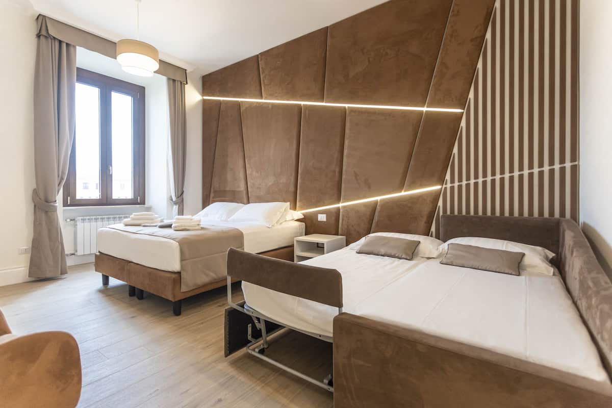 A modern hotel room with two beds, a large window with curtains, a wooden floor, and a wall-mounted light fixture.