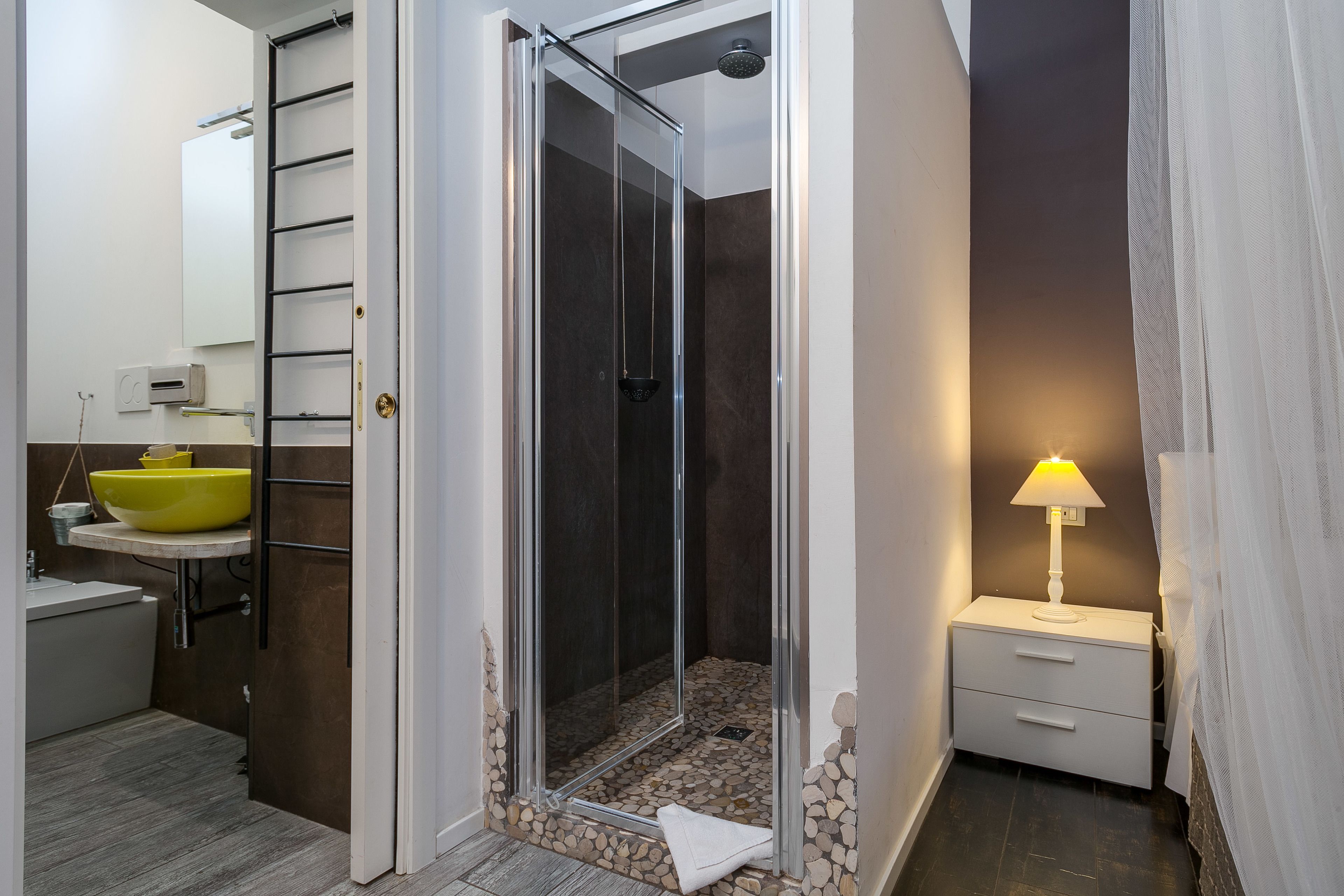 A modern bathroom with a glass-enclosed shower, a yellow sink, and a bedside table with a lamp.