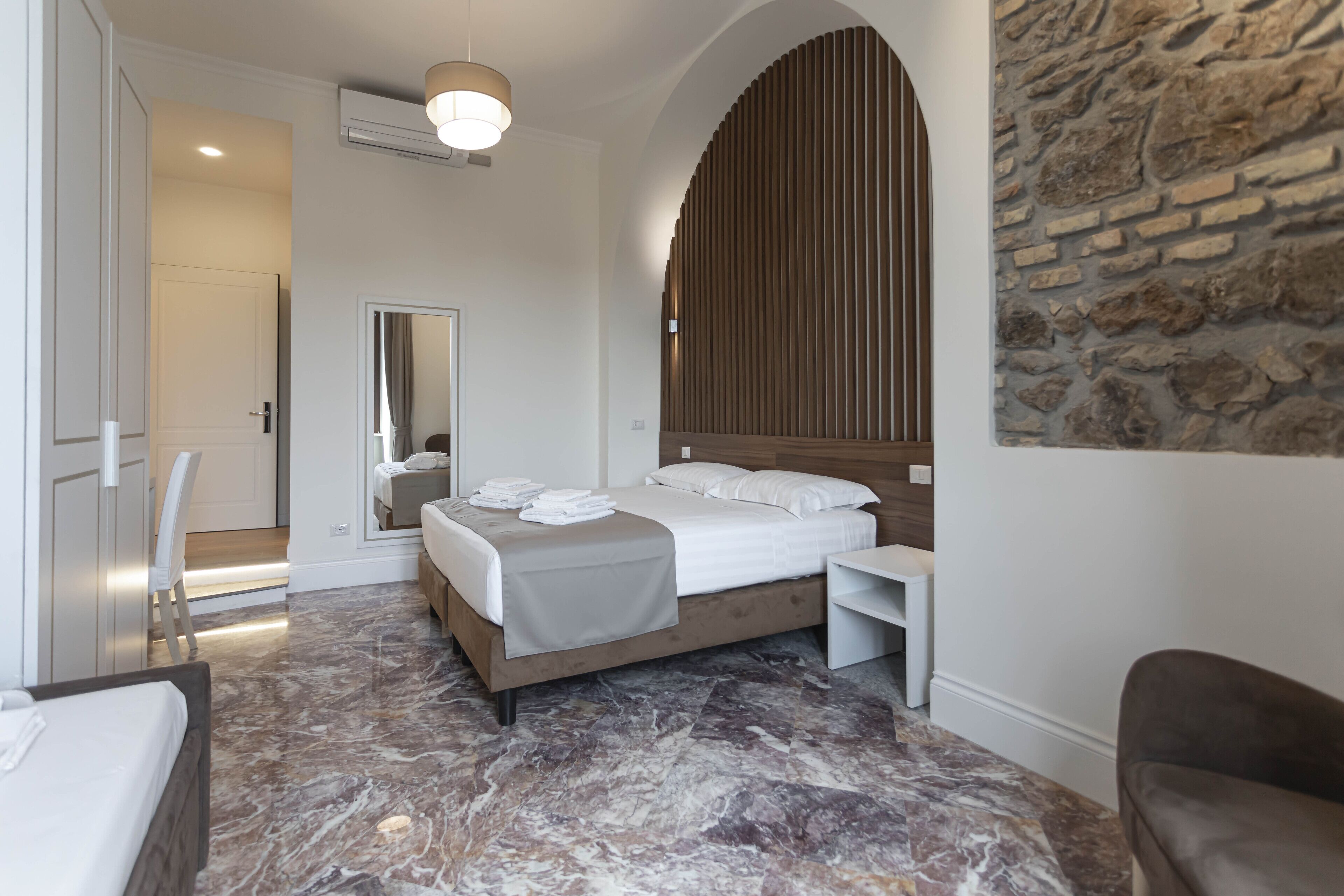 Habitació d'hotel moderna amb sòl de marbre, un llit gran amb capçalera de fusta, una taula de nit, una cadira i una paret decorativa de pedra.