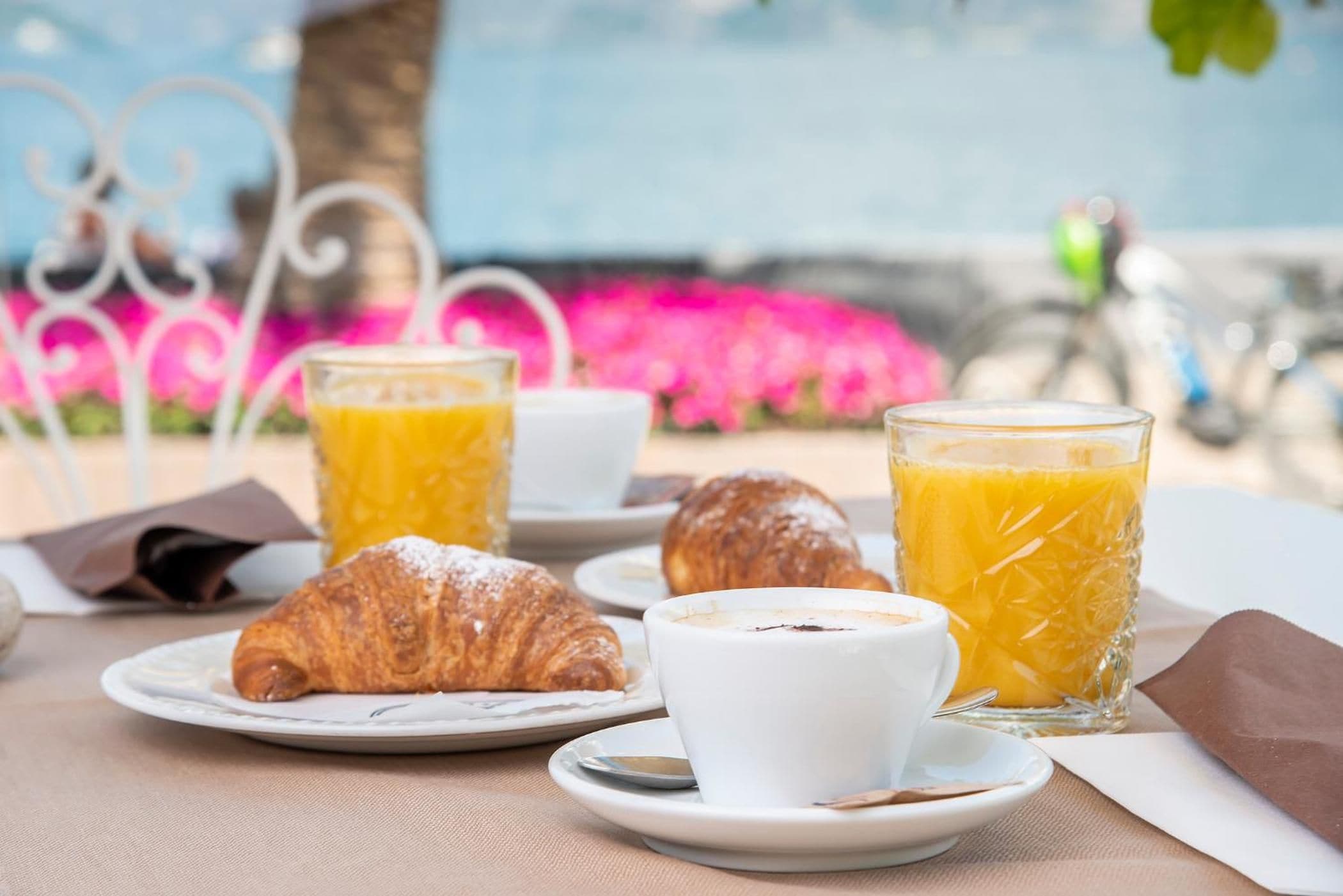 Une table dressée pour le petit-déjeuner, avec des croissants, du café et du jus d’orange, et en toile de fond, une vue sur des fleurs et une clôture en métal.