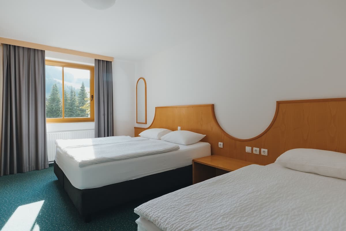 Ein Hotelzimmer mit zwei Betten, einem Holzkopfteil, einem Fenster mit Vorhängen und Blick auf Bäume.