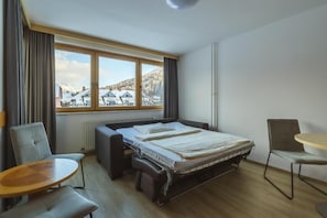 Habitación de hotel con cama, dos sillas, una mesita y ventana con vistas a casas y montañas.