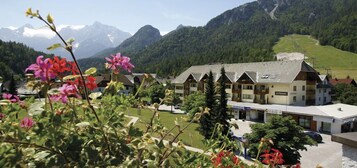 A hotel with a traditional Alpine architectural style, featuring a central building with multiple windows and a sloped roof, surrounded by lush greenery and mountains in the background.