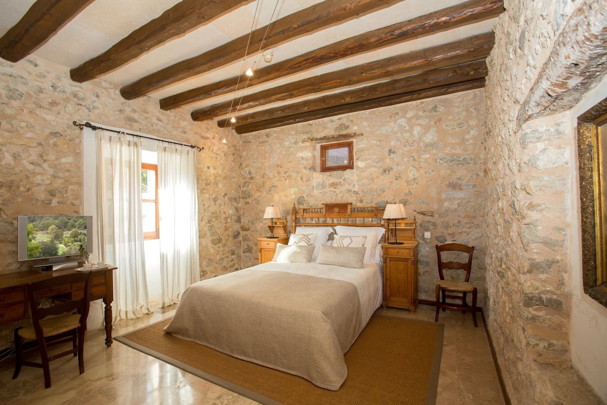 A bedroom with stone walls, a wooden bed, a desk with a television, and a chair.