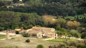 Un edificio de piedra tradicional con tejado de tejas, rodeado de exuberante vegetación y un paisaje montañoso.