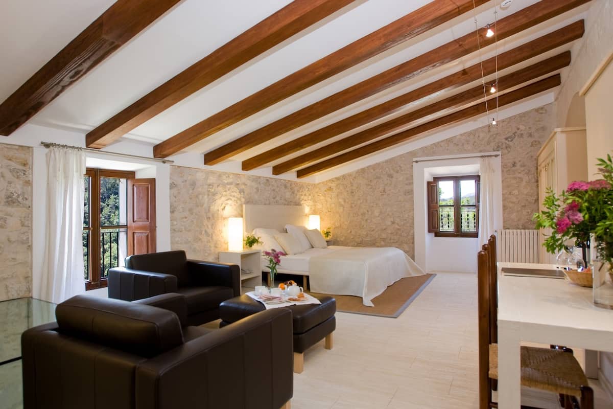 A spacious room with a high ceiling and exposed wooden beams, a bed with white linens, a seating area with leather sofas, and a dining table with a vase of flowers.
