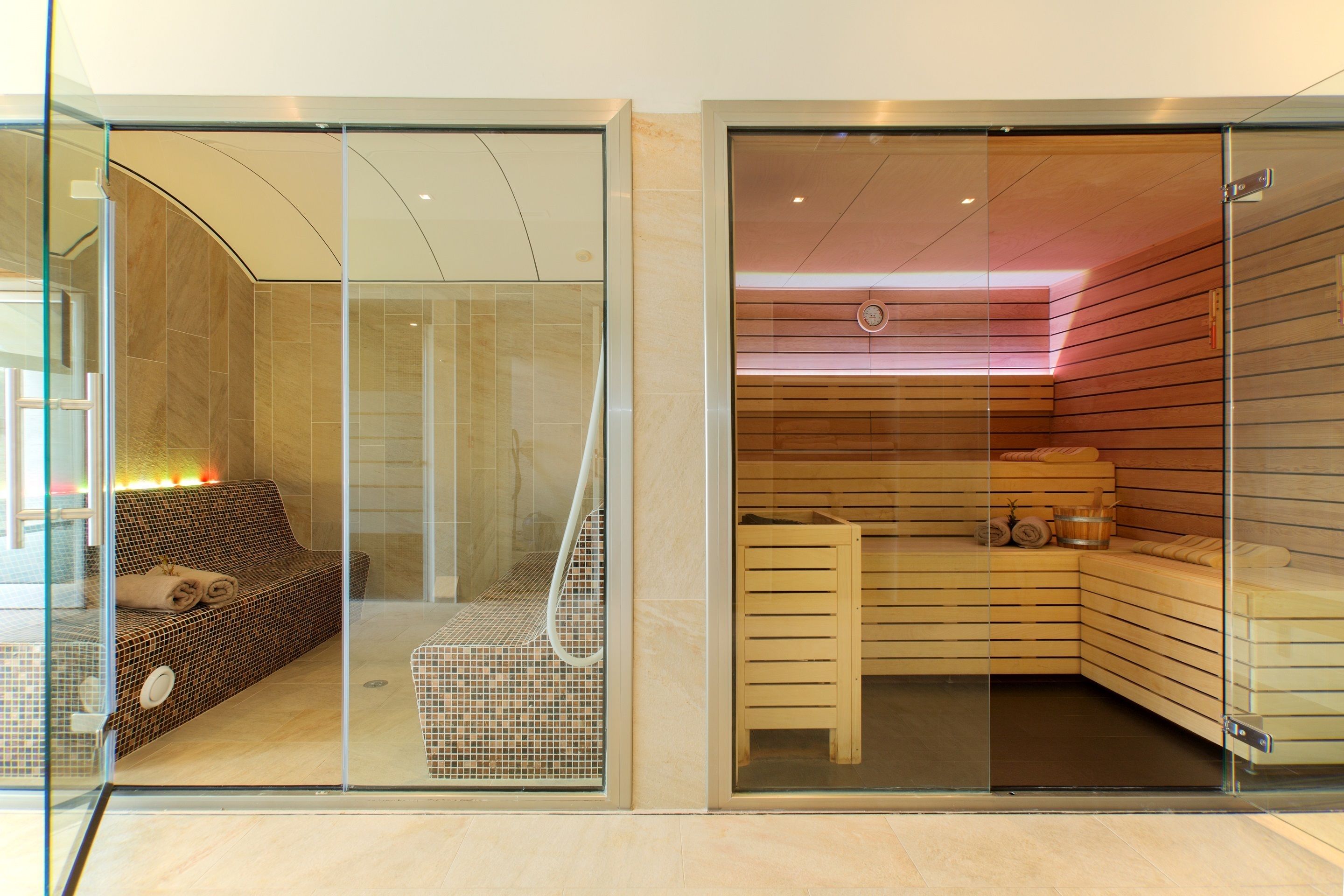 A modern spa room with a glass partition, a wooden sauna, and a relaxation area with a sofa and towels.