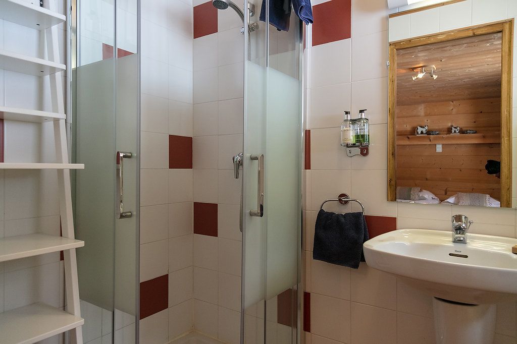 A bathroom with a glass-enclosed shower, white tiles with red accents, a white sink, and a wooden shelf with toiletries.