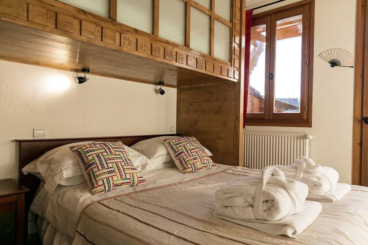 A bedroom with a wooden headboard, two pillows with a geometric pattern, a neatly made bed with white linens, and a window with a view of the outside.