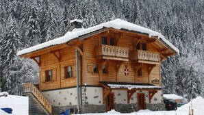 A wooden chalet with a balcony, a stone foundation, and a snow-covered roof, surrounded by a forest. - Chilly Powder - Hôtel au Coin du Feu (Morzine)