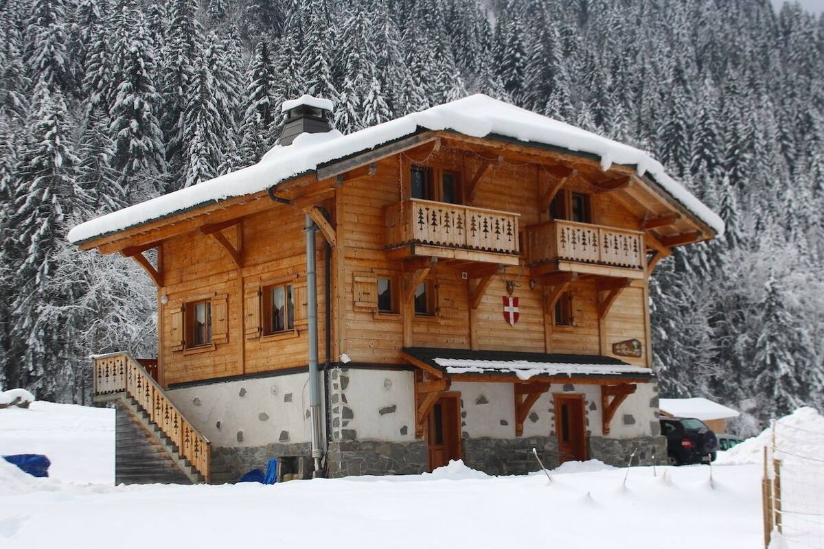 A wooden chalet with a balcony, a stone foundation, and a snow-covered roof, surrounded by a forest.