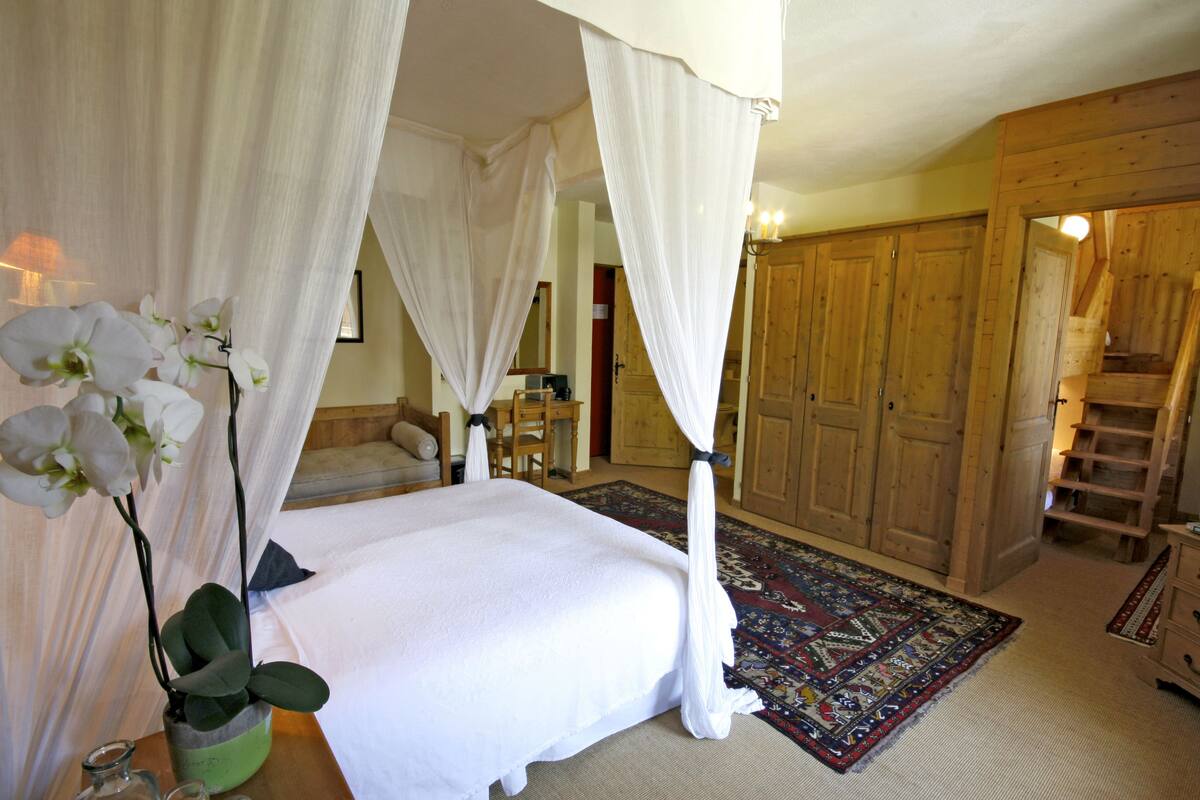A bedroom with a four-poster bed, a large window, wooden wardrobe, and a small table with a vase of flowers.