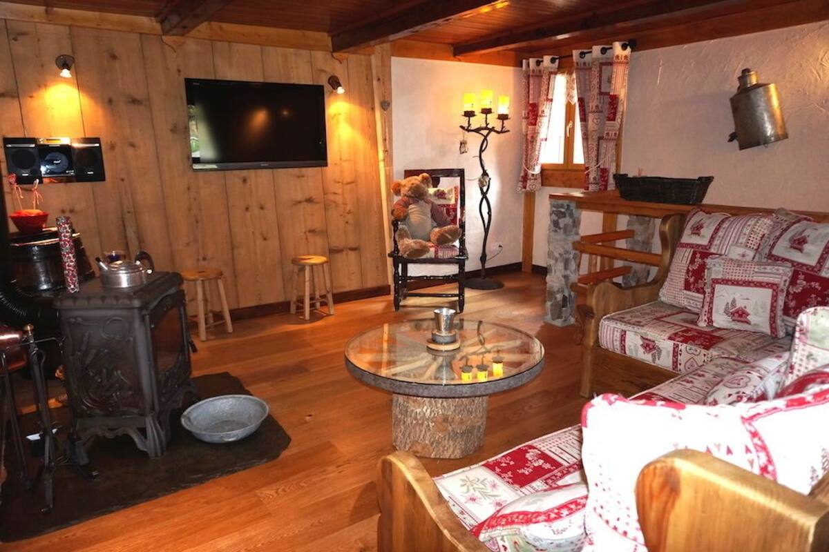 A cozy room with a wooden stove, a round glass table, a sofa with patterned cushions, a television, and a wall-mounted lamp.