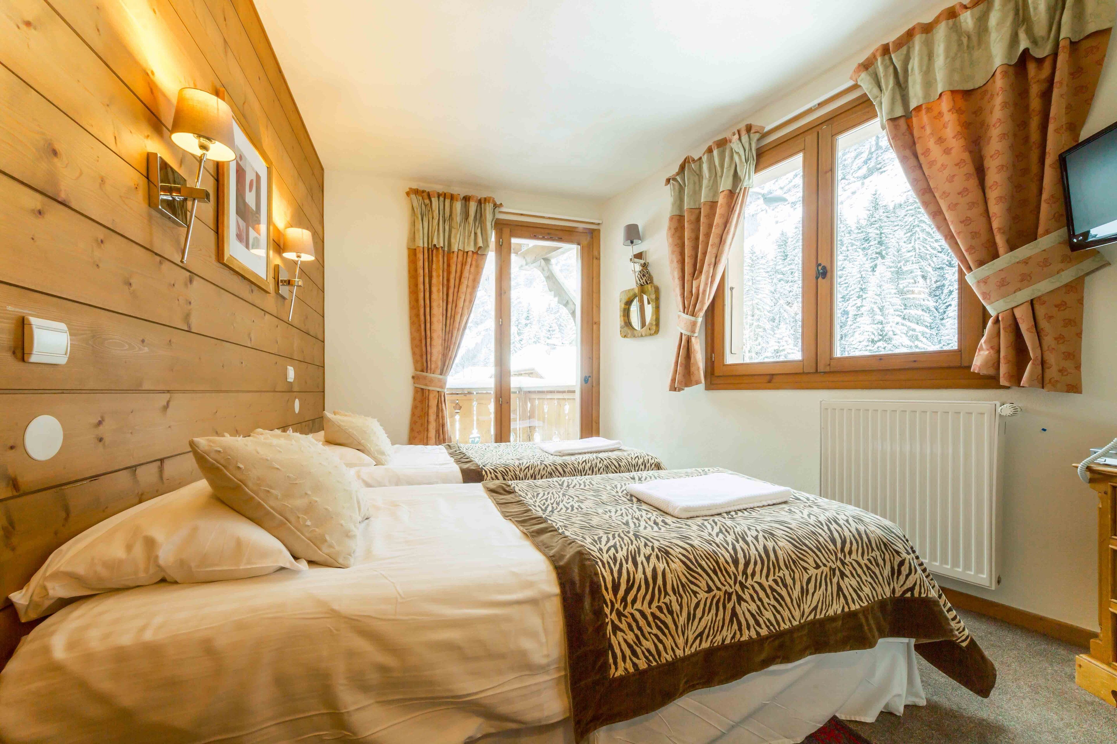 A hotel room with two beds, wooden paneling, a television, and a view of trees through the window.
