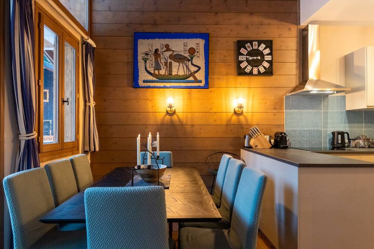 A wooden interior with a dining table, blue upholstered chairs, a wall-mounted clock, and a framed picture.