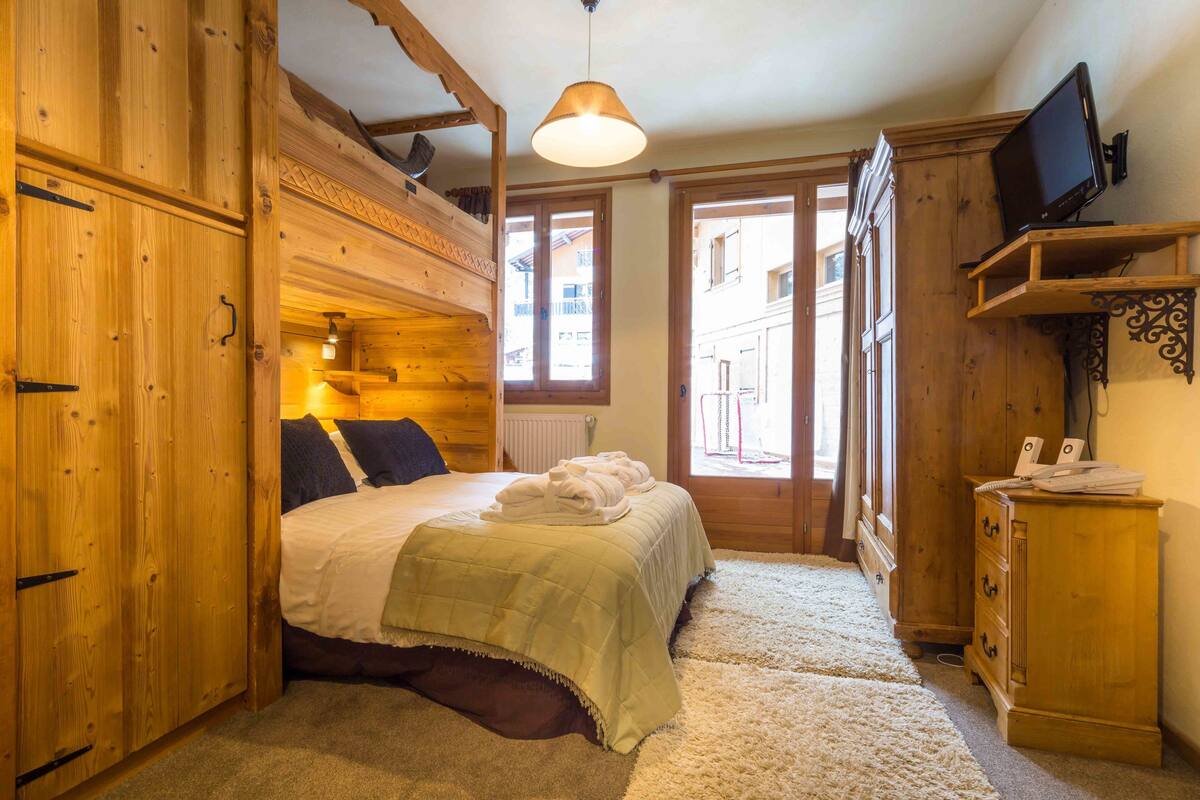 A cozy wooden room with a bed, a television on a shelf, and a view through a door.