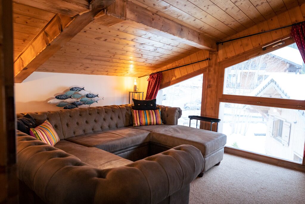 A cozy room with a large sectional sofa, a wooden ceiling, and a window with a view of buildings outside.