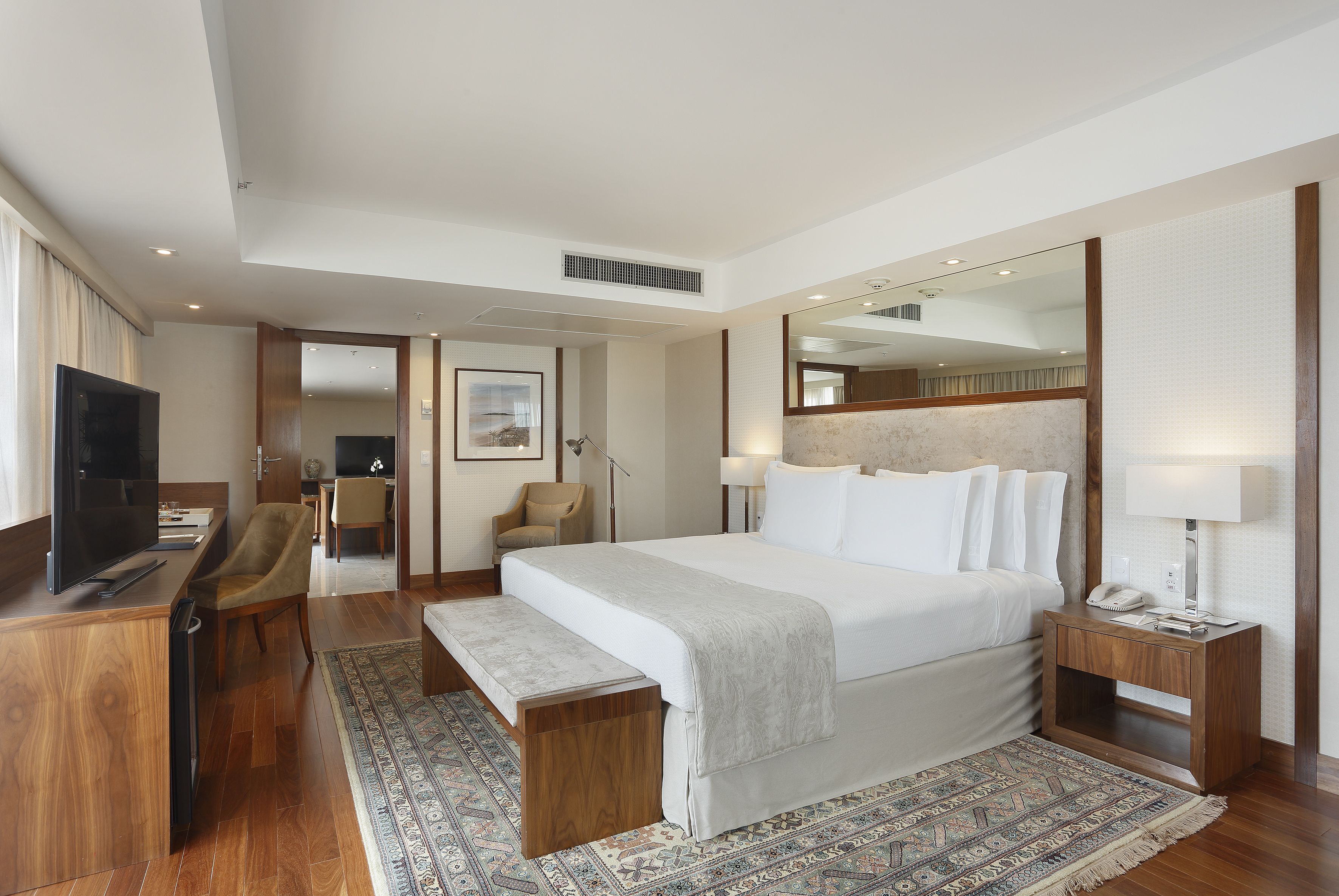 A hotel room with a large bed, bedside tables, a desk with a TV, and a patterned rug.