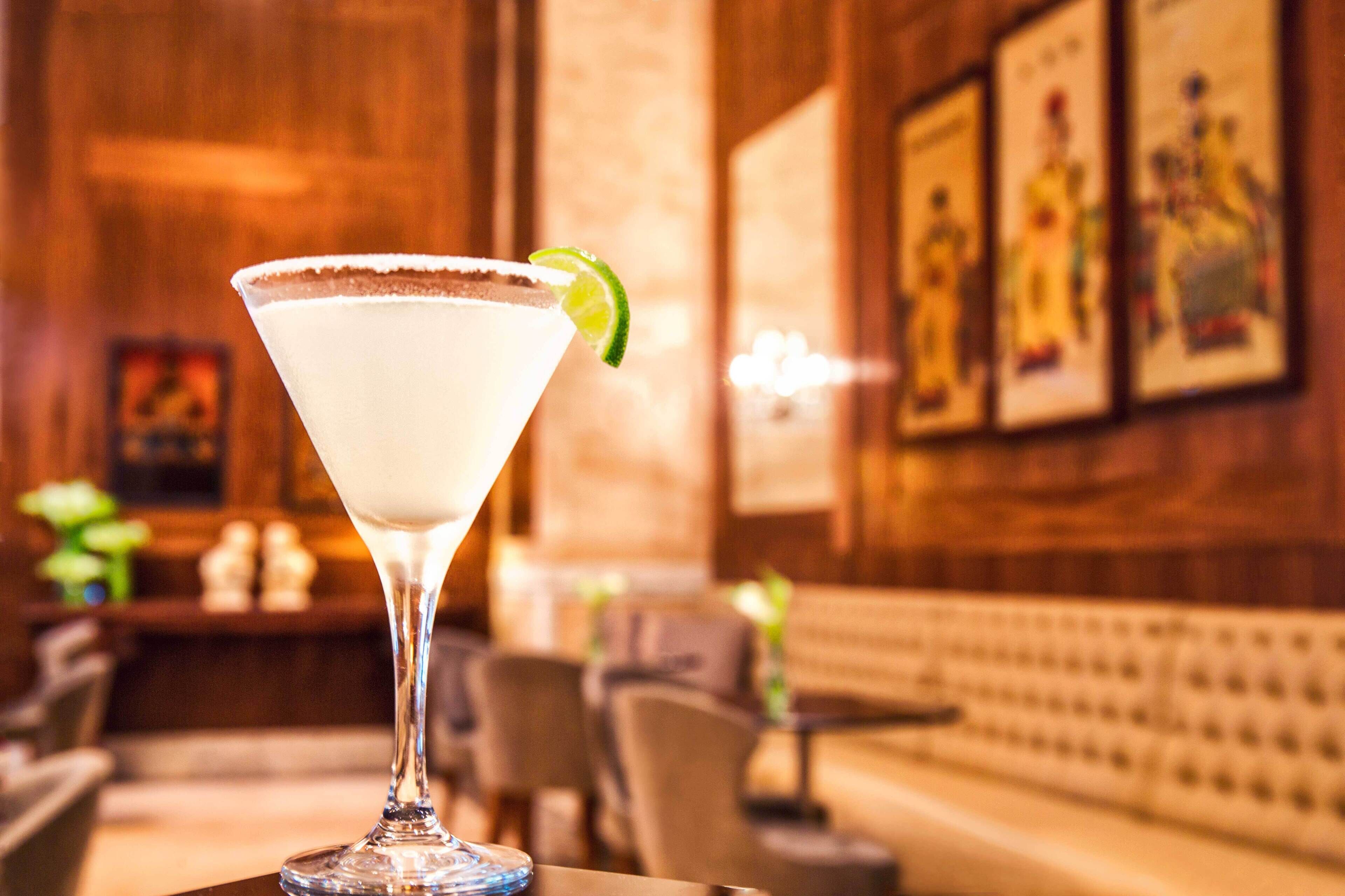 Una copa de martini con un adorno de lima sobre una barra en una habitación cálidamente iluminada, con decoración de madera y cuadros en la pared.