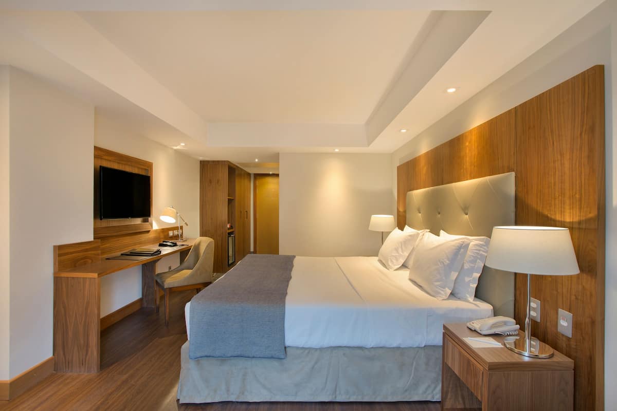 Ett modernt hotellrum med en stor säng, ett skrivbord med en stol, en tv och ett nattduksbord med en lampa.