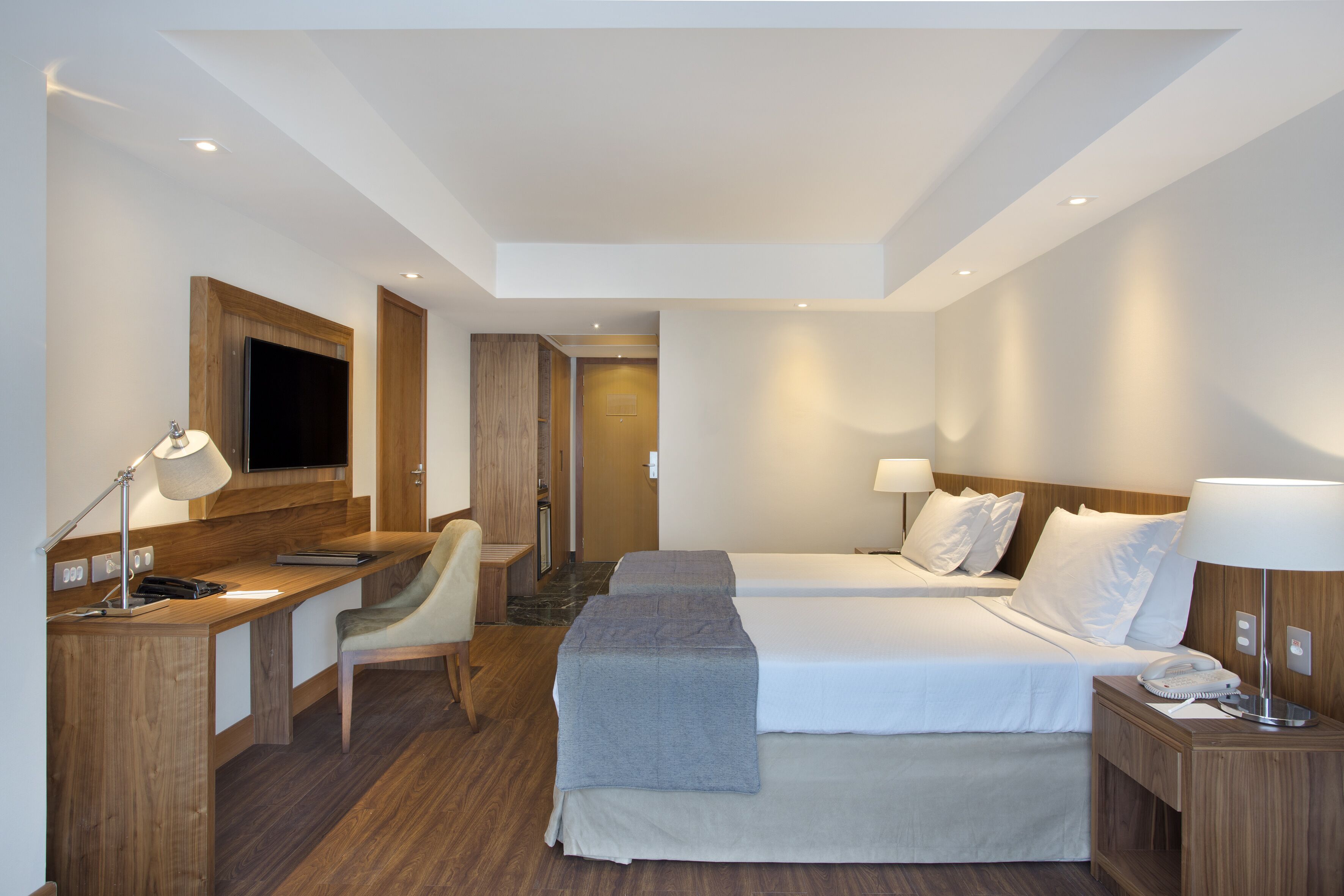 A hotel room with two beds, a desk, a chair, and a TV.