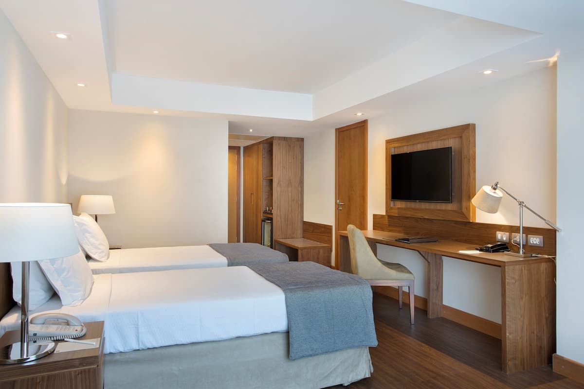 Ett modernt hotellrum med två sängar, ett skrivbord, en stol, en TV och en lampa.
