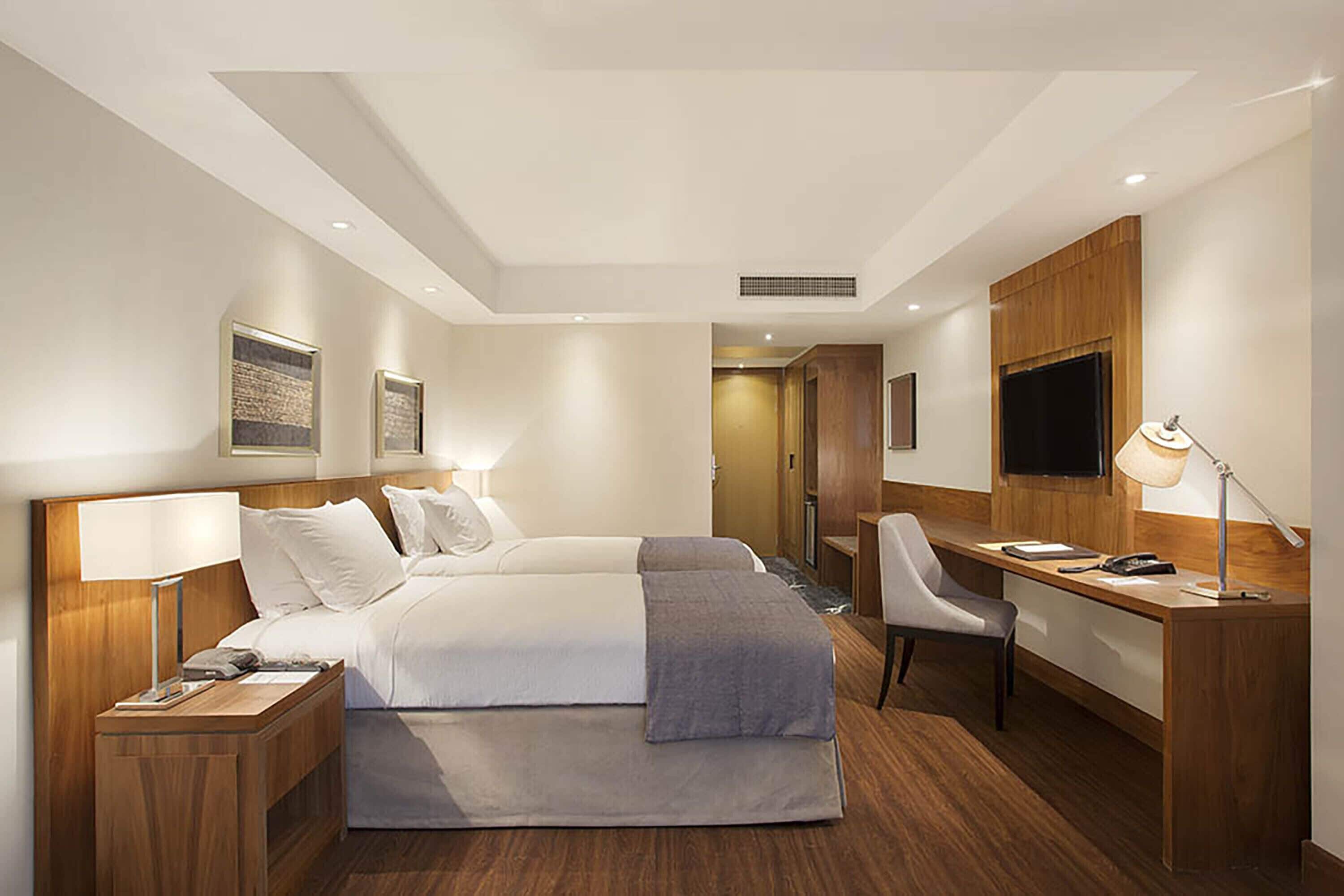 A modern hotel room with two beds, a desk, a chair, a TV, and a lamp.
