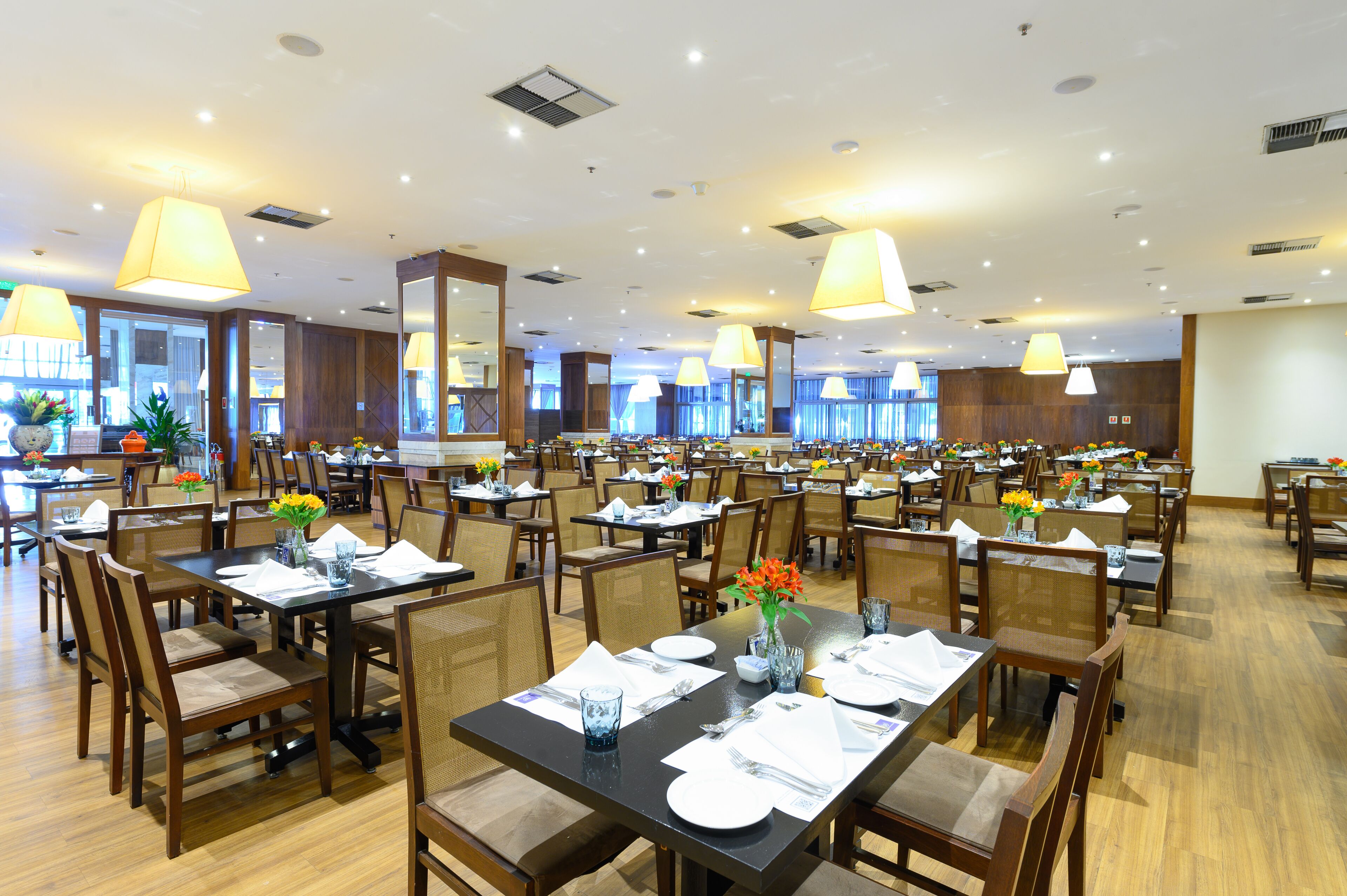 A spacious dining area with multiple tables and chairs, large hanging lamps, and potted plants.