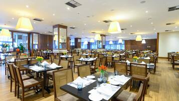 A spacious dining area with multiple tables and chairs, large hanging lamps, and potted plants.