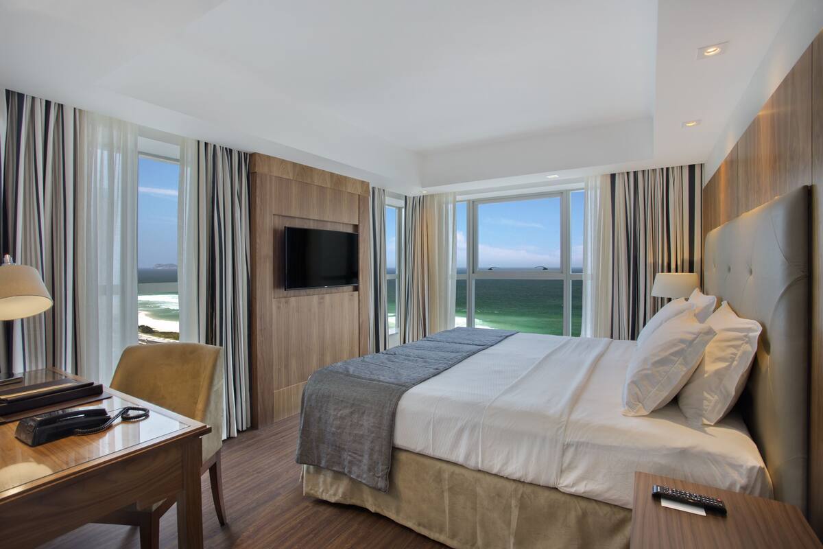 Ett hotellrum med en stor säng, ett skrivbord med en lampa, en TV och utsikt över havet.