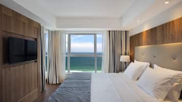 A hotel room with a large bed, a flat-screen TV mounted on the wall, and a view of the ocean through a large window.
