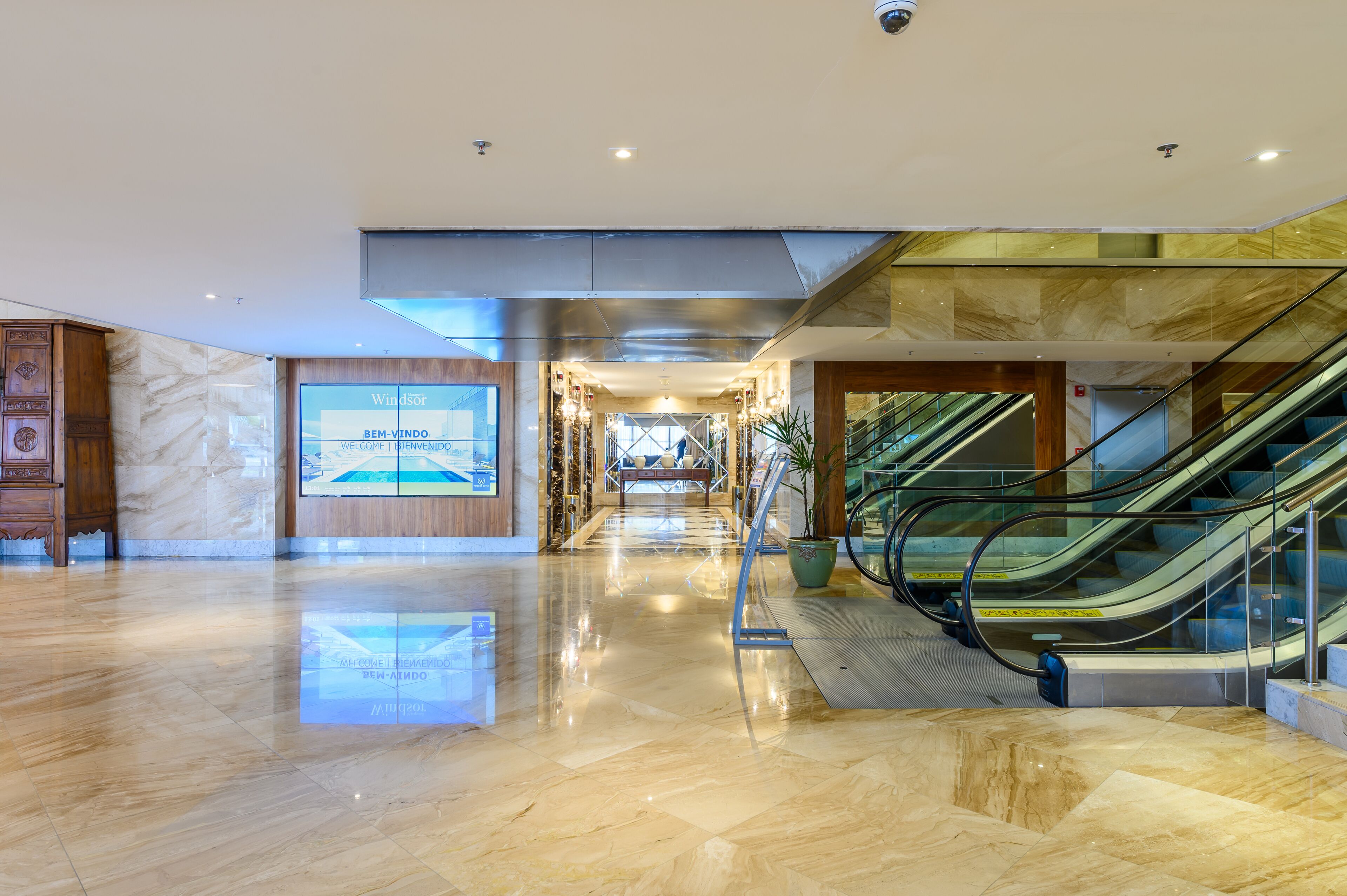 A modern hotel lobby with a marble floor, a large digital screen displaying 'Windsor', and a staircase with glass railings.