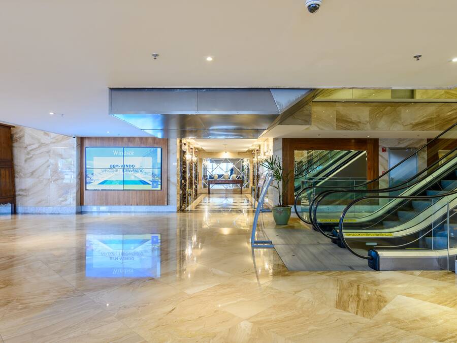 Un moderno vestíbulo de hotel con piso de mármol, una gran pantalla digital mostrando 'Windsor' y una escalera con barandas de vidrio.